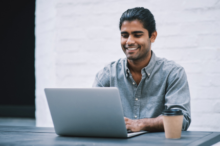 A man with his hair tied back, working from a laptop on an outdoor table