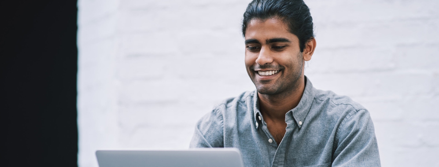 A man with his hair tied back, sitting at an outdoor table on a laptop