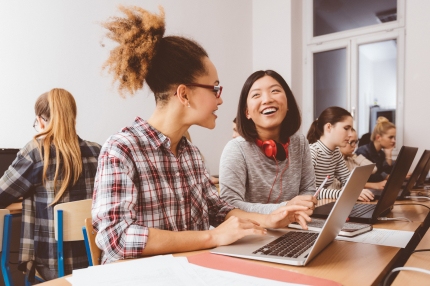 Two female students sitting at a work desk with laptops and smiling at each other, with other students working in the background