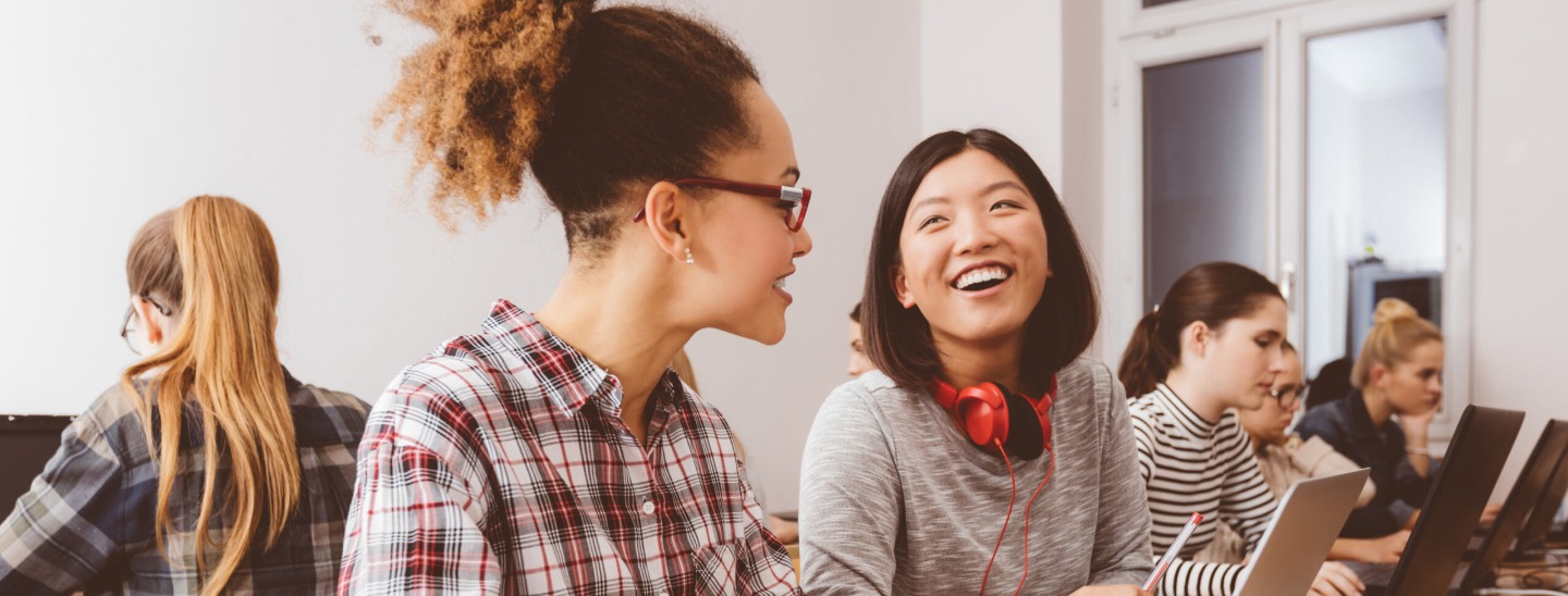 Two female students sitting beside each other and smiling, sitting amongst other students on laptop computers