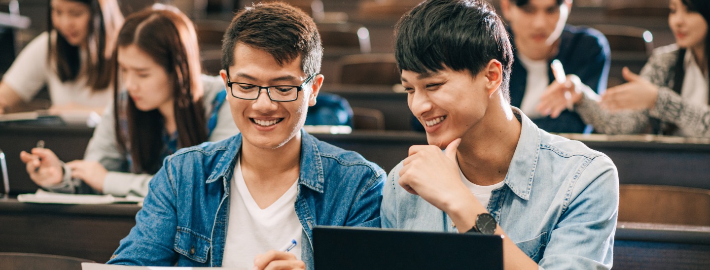 Two students sitting together in a lecture hall, looking at a piece of paper in one of their hands