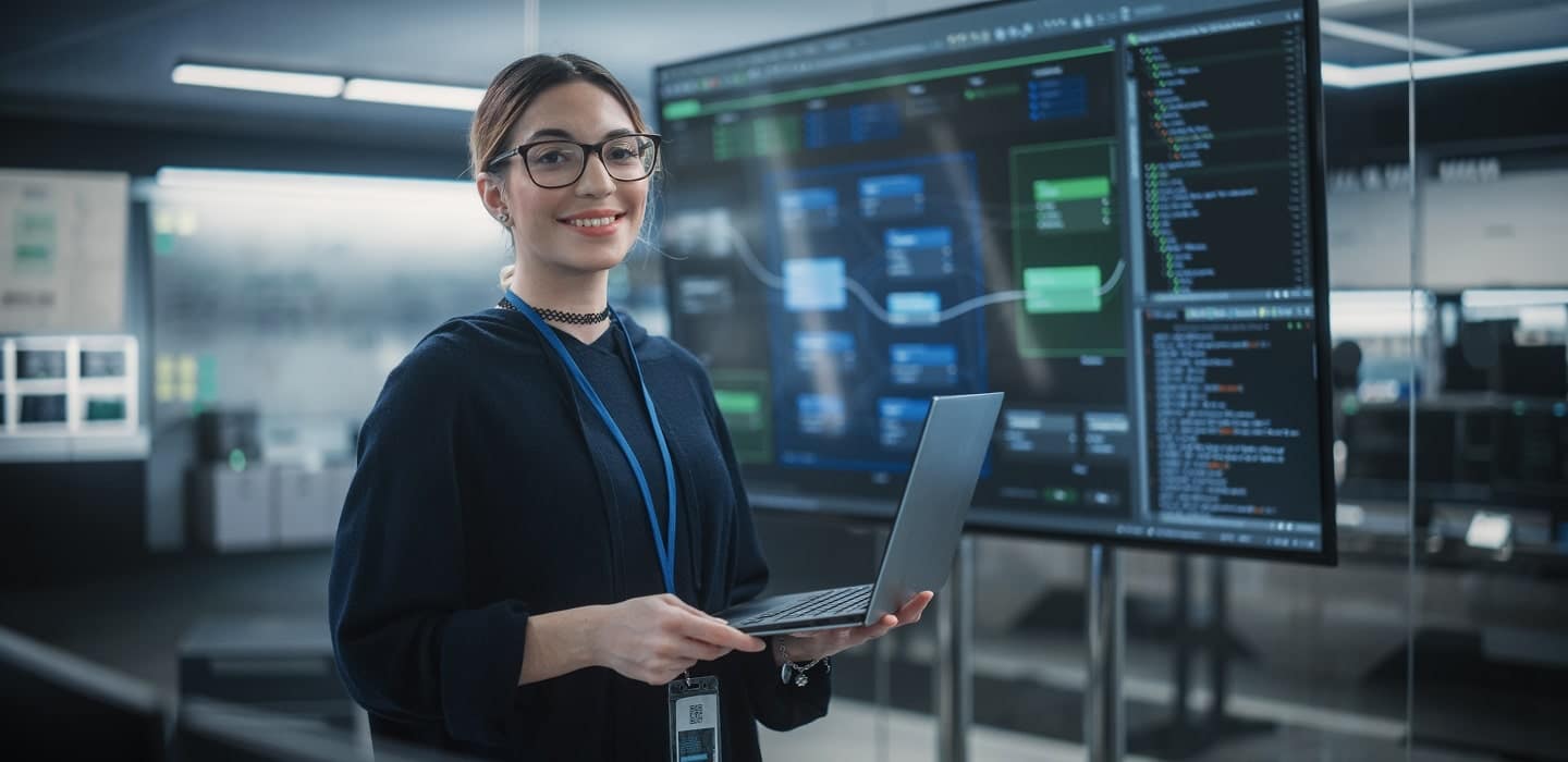 A smiling woman with glasses holding a laptop, standing in front of a large tv monitor