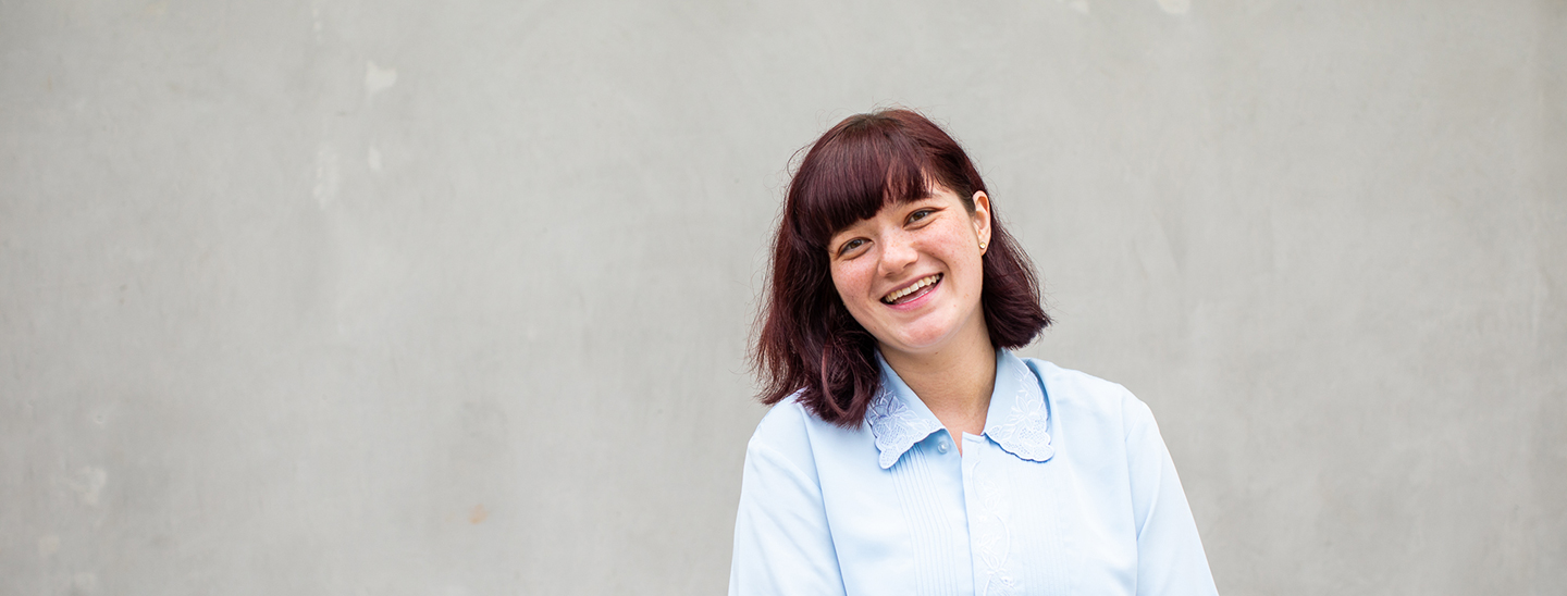 A young female international student with red hair and a a light blue blouse, smiling towards camera (1440x548).