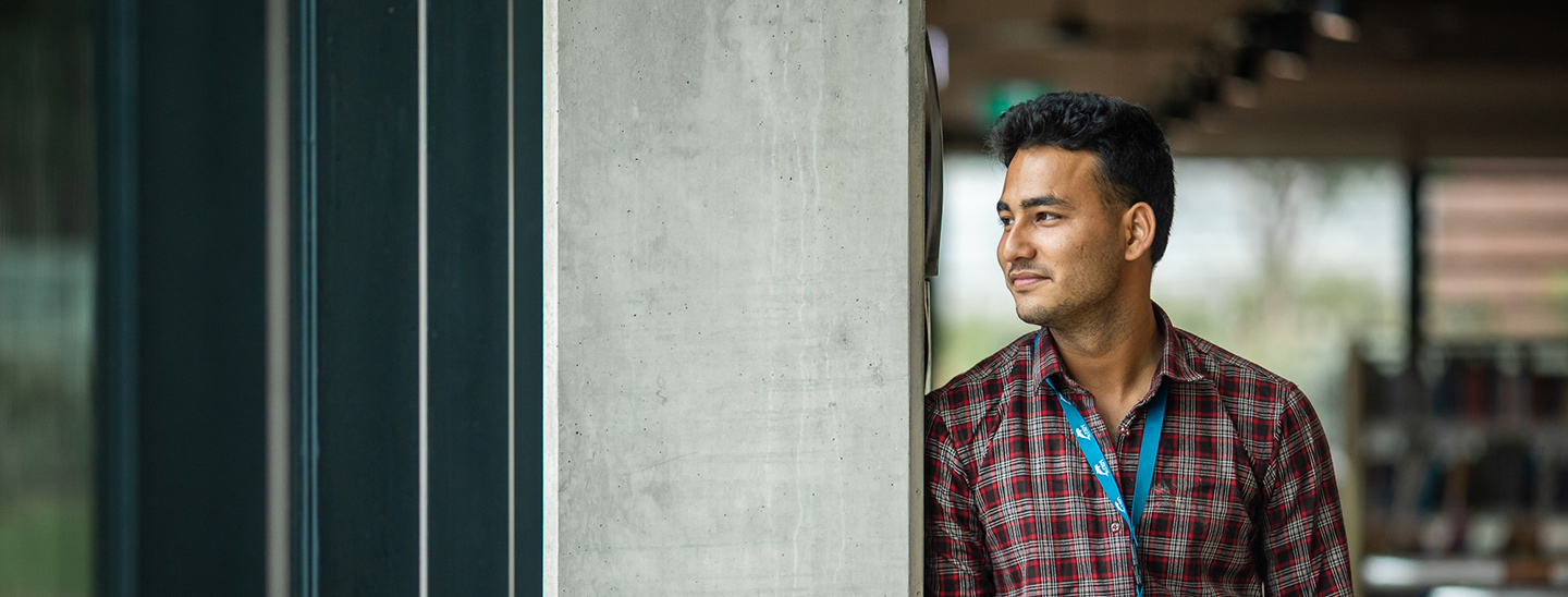A male international student wearing a checkered shirt and a lanyard, standing with his hands in his pockets and looking to his right side (1440x548).