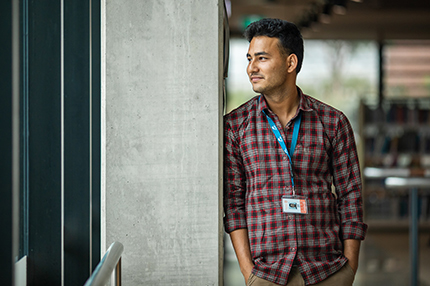 A male international student wearing a checkered shirt and a lanyard, standing with his hands in his pockets and looking to his right side (430x286).