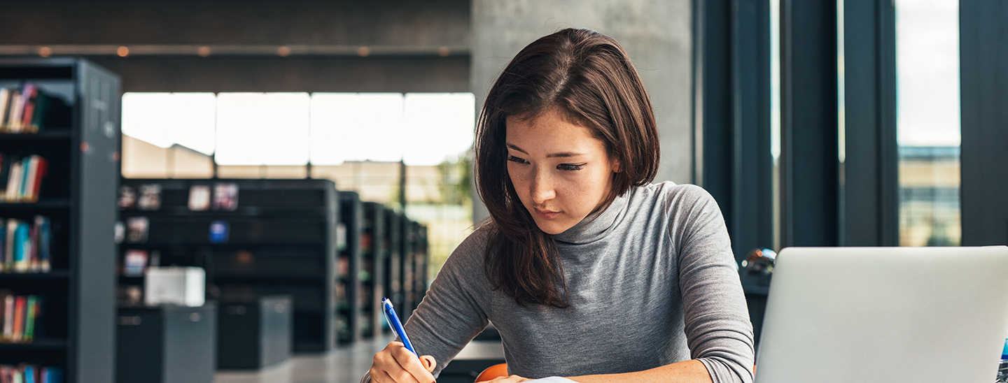 A young female Bachelor of Accounting student working with a pen and notebook, with an open laptop in front of her (1440x548)