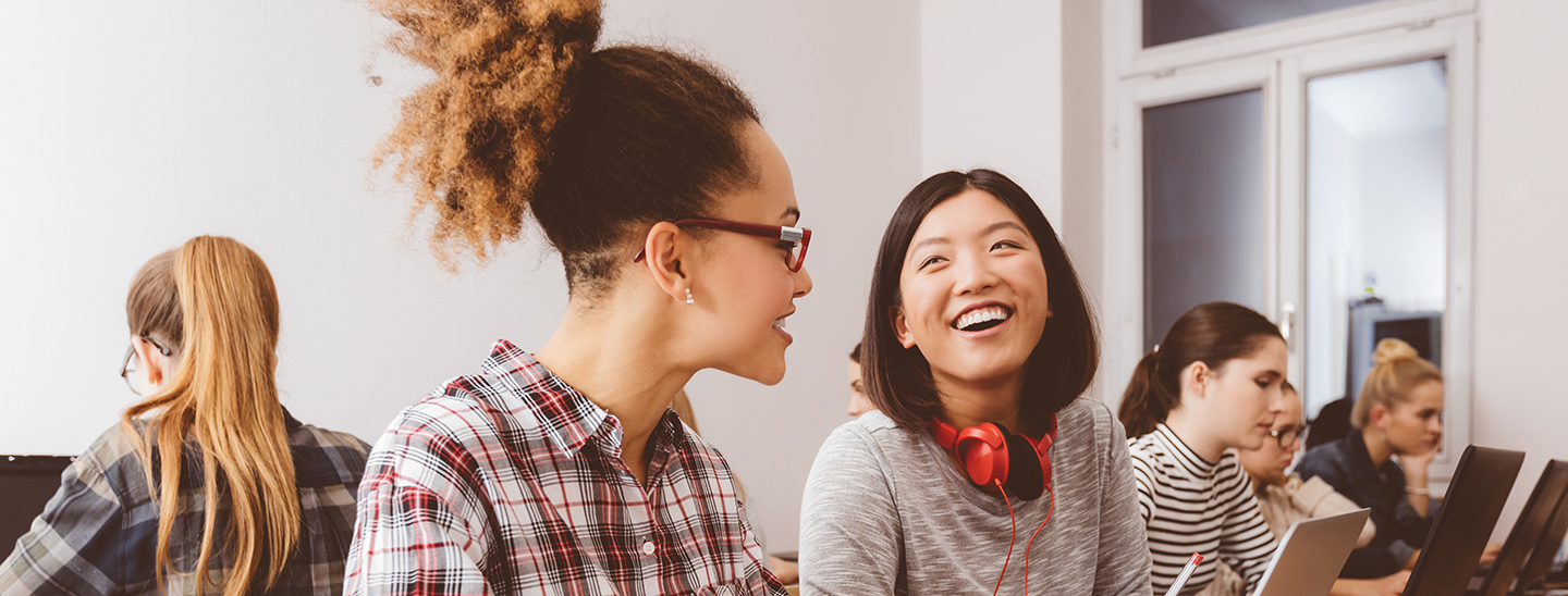 Two young female Bachelor of Business students sitting together at a long desk with other students, using laptops and smiling towards each other (1440x548)