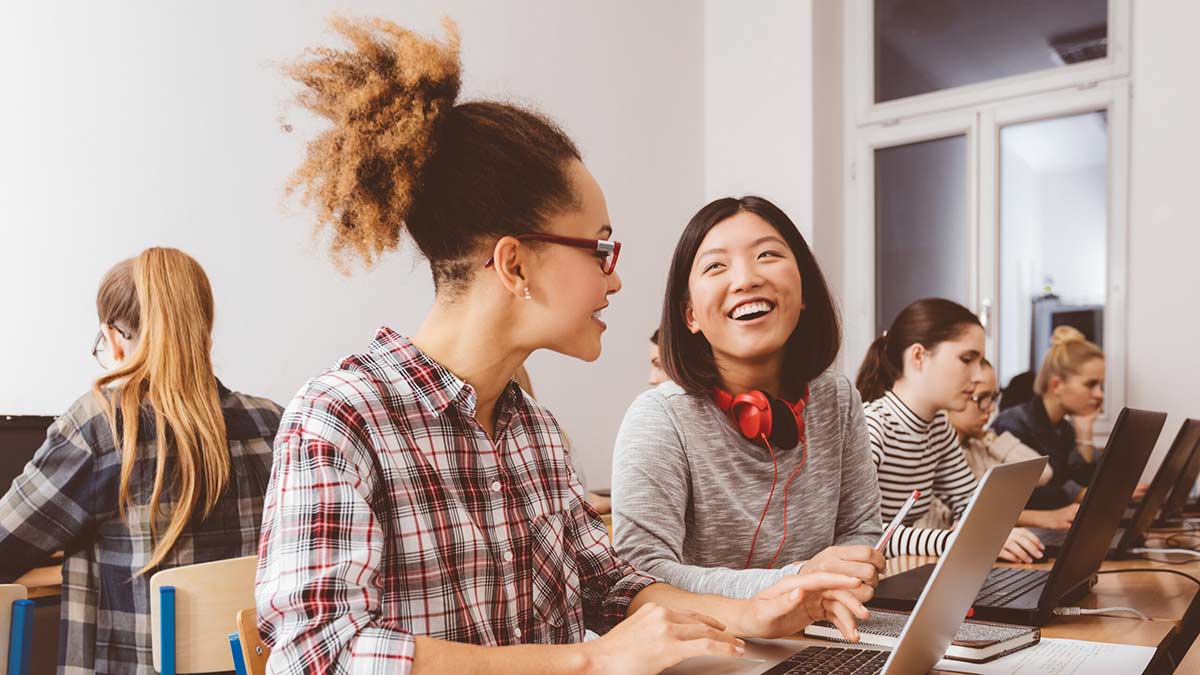 Two young female Bachelor of Business students sitting together at a long desk with other students, using laptops and smiling towards each other (1200x675)