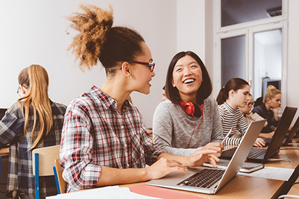 Two young female Bachelor of Business students sitting together at a long desk with other students, using laptops and smiling towards each other (430x286)