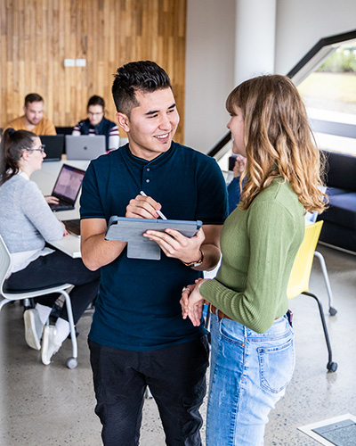 Two Bachelor of Business students standing together in a campus common area, with one student holding a stylus and tablet (400x500).