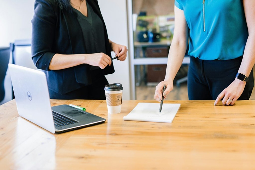 Photo of a desk with an open laptop, coffee and piece of paper, with the torsos of two women facing each other