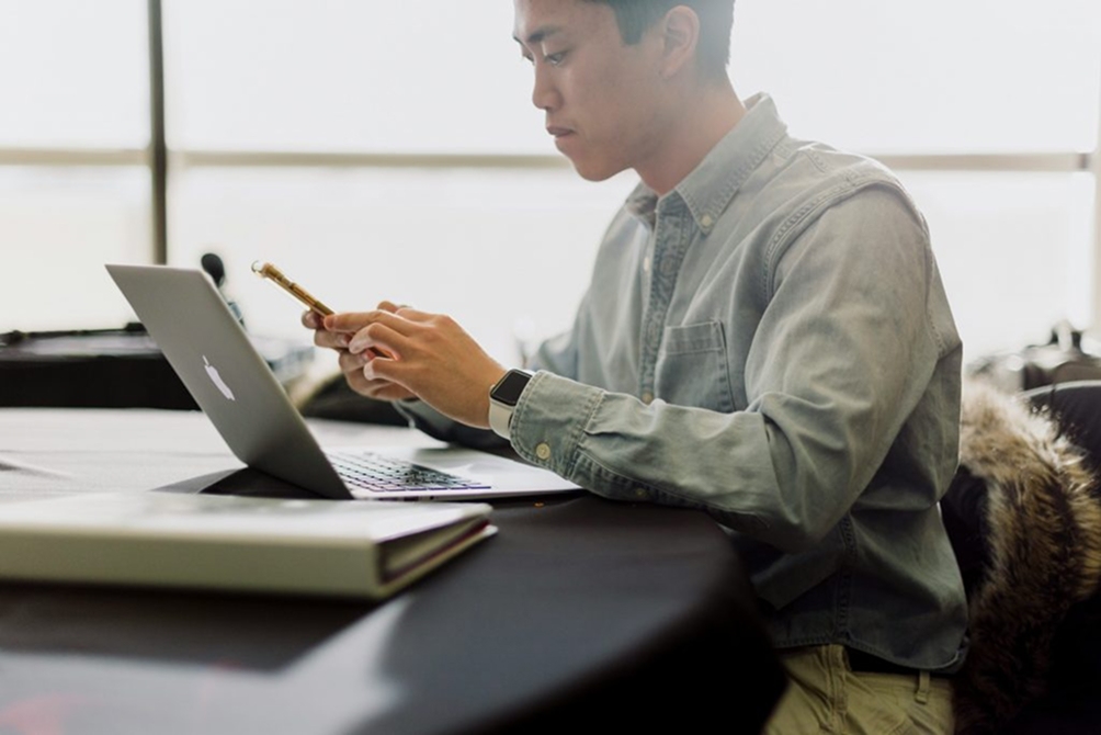 A man in a collared shirt sitting at a desk in front of a laptop, looking at a mobile phone in his hands