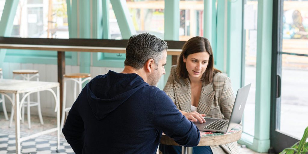 A woman and man sitting at a high table in a restaurant, both looking towards a laptop screen - Certified Practising Accountant (CPA)