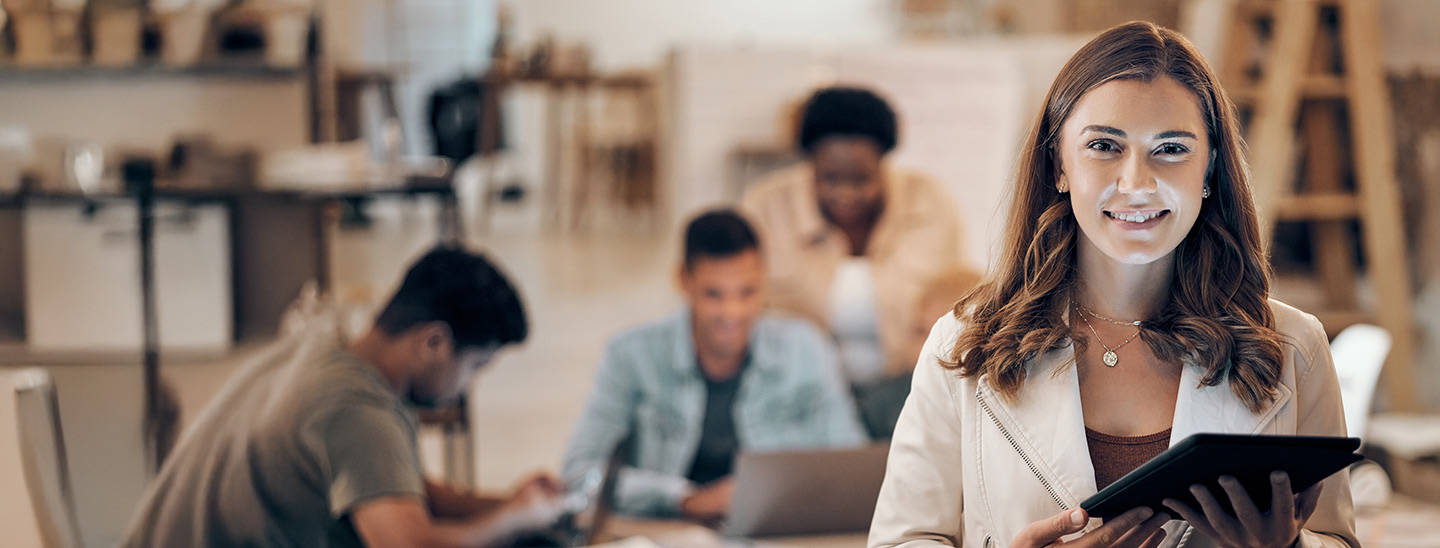 A woman standing in an office and holding a tablet in her hand, with her colleagues working at a desk in the background (1440x548)