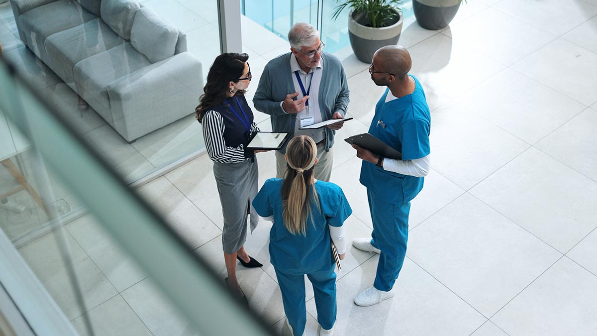 Four health professionals standing in a circle in a hospital common area, with two wearing professional business attire and two wearing scrubs (medical/surgical attire) - 1200x675