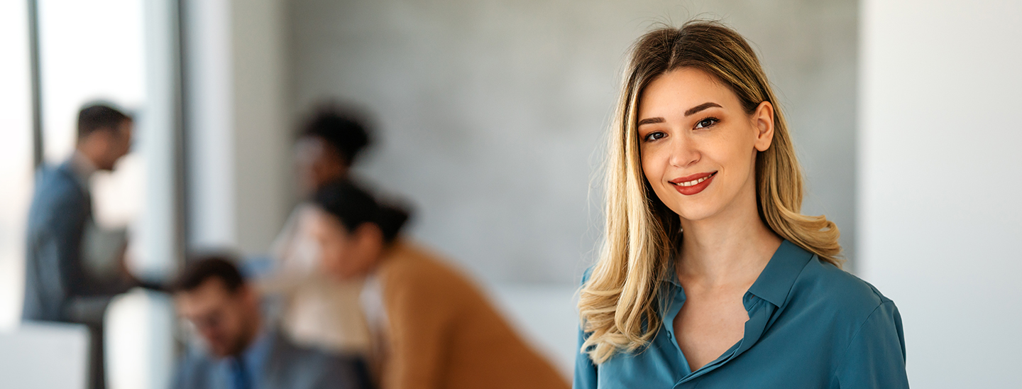 A female Master of Health (Professional Practice) student standing in an office space with colleagues working at desks in the background (1440x548)