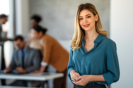 A female Master of Health (Professional Practice) student standing in an office space with colleagues working at desks in the background (430x286)