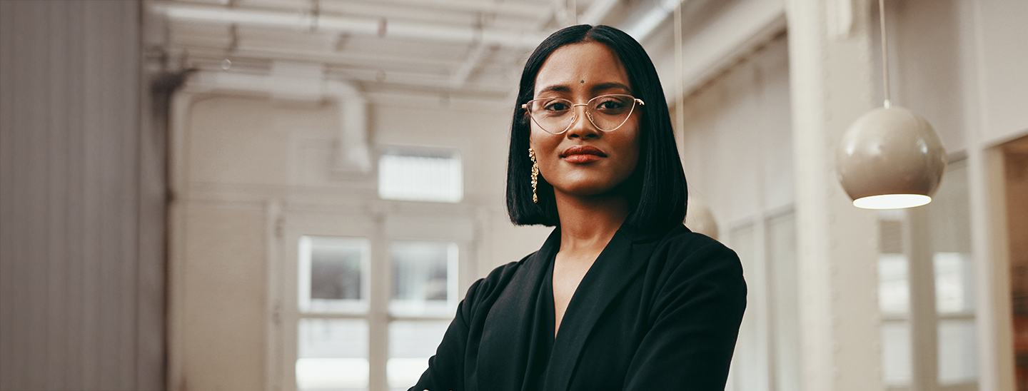 A female Master of Professional Accounting student wearing glasses and standing with her arms folded in a classroom (1440x548)
