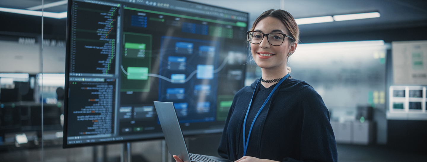 A female Master of Professional Information Technology student wearing glasses and standing with a laptop in her hands (1440x548)