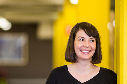 A female student leaning against a yellow wall and smiling - Master of Professional Accounting (Professional Practice)