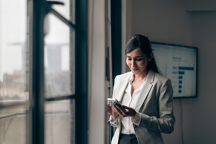 A female Master of Project Management and Leadership (Professional Practice) student standing in a room in front of a large window, looking downwards at a mobile phone in her hand