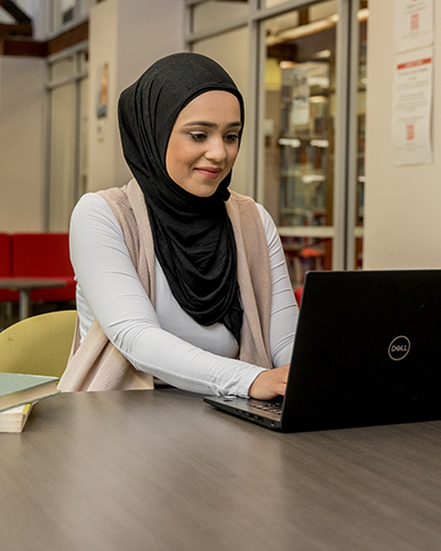 A female Master of Professional Accounting student sitting in a campus common area and working on a laptop (400x500).