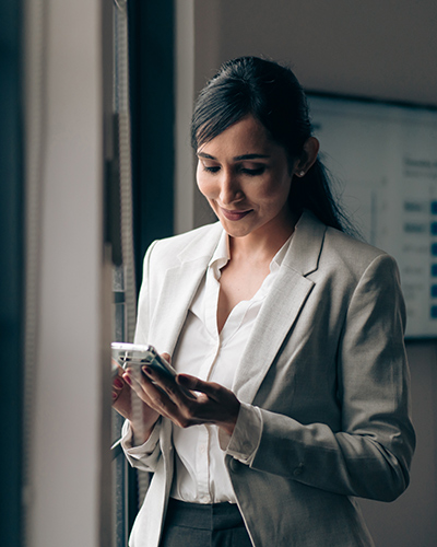 A female Master of Project Management and Leadership student standing in an office space, looking downwards towards a smartphone in her hand (400x500)