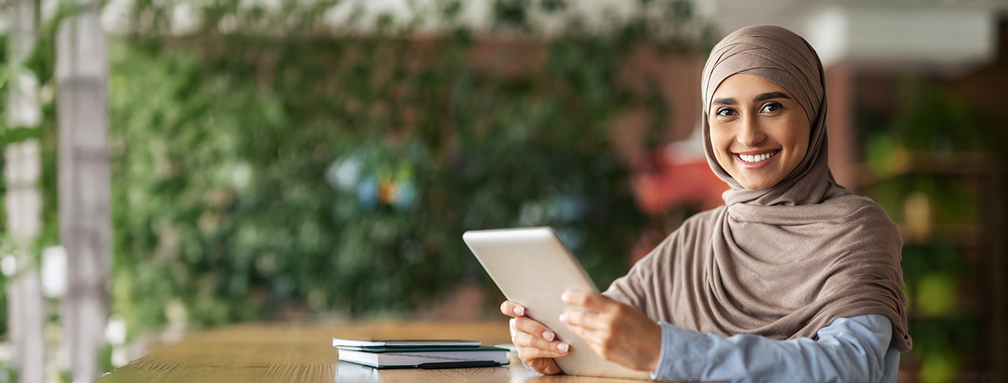 A woman sitting at a long desk with a tablet in her hands and a stack on notebooks in the background (1440x548)