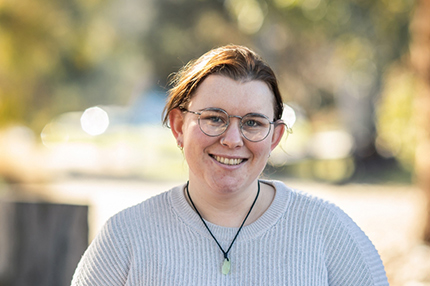 A young woman with glasses smiling to camera, standing in an outdoor location with the background out of focus