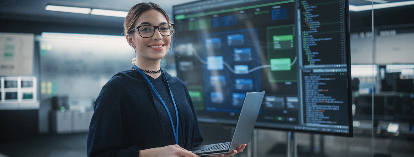 A smiling woman with glasses holding a laptop, standing in front of a large tv monitor