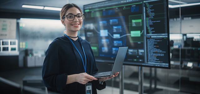 A smiling woman with glasses holding a laptop, standing in front of a large tv monitor