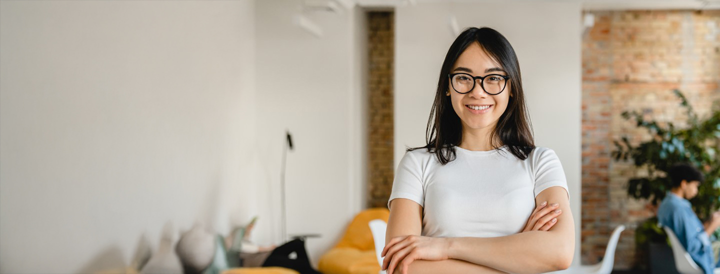 A female student wearing glasses and standing with her arms folded in an office space - 1440x548