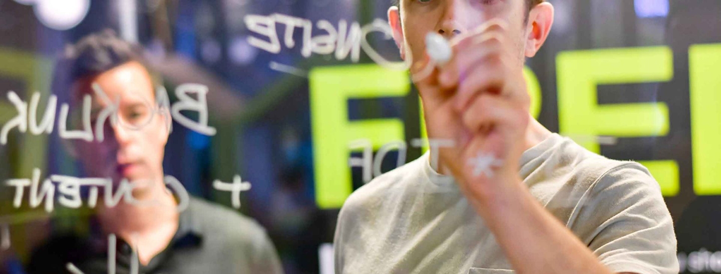 A man writing on a clear screen with a marker pen, with a man in the background watching over his shoulder