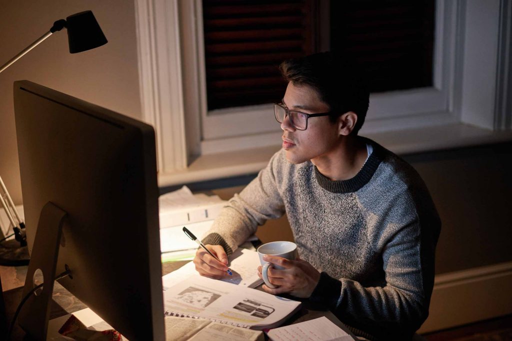 A young male student wearing glasses and holding a coffee mug and pen in his hands, working at a desk on an iMac computer monitor - Data Analytics