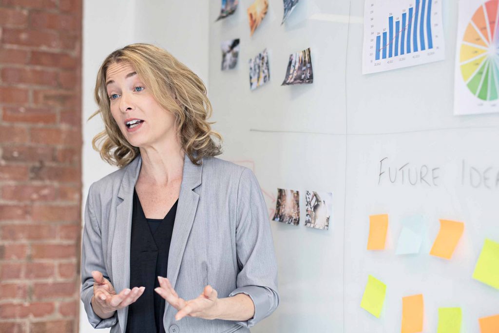 A female teacher with short blonde hair standing in front of a whiteboard and gesturing with her hands - Data Analytics